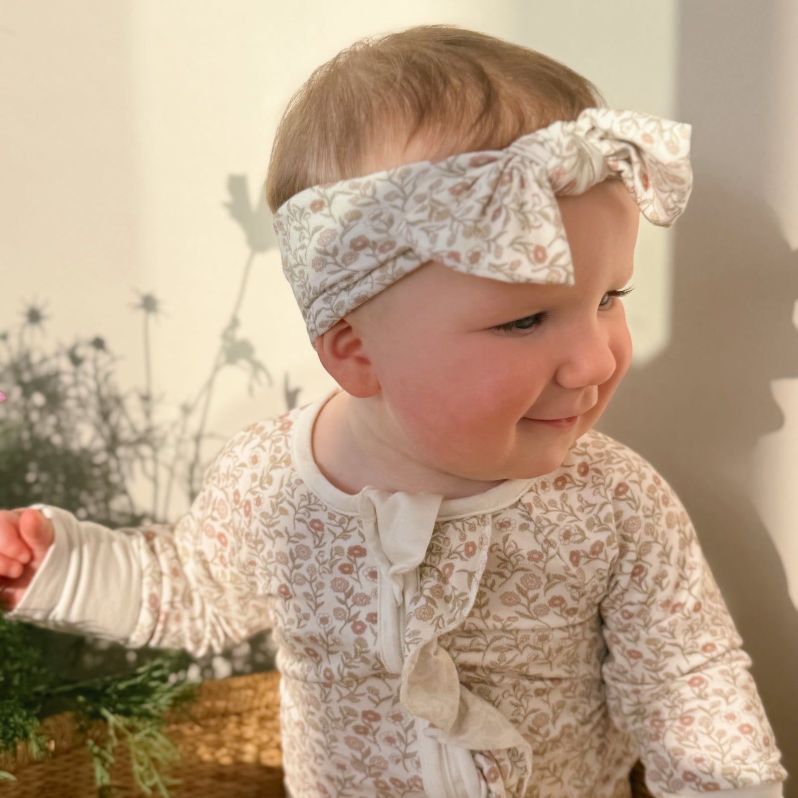 Baby wearing a floral outfit and headband, standing outdoors with plants in the background