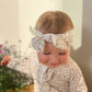 Baby wearing a floral headband and matching outfit, sitting on a couch with a blurred background