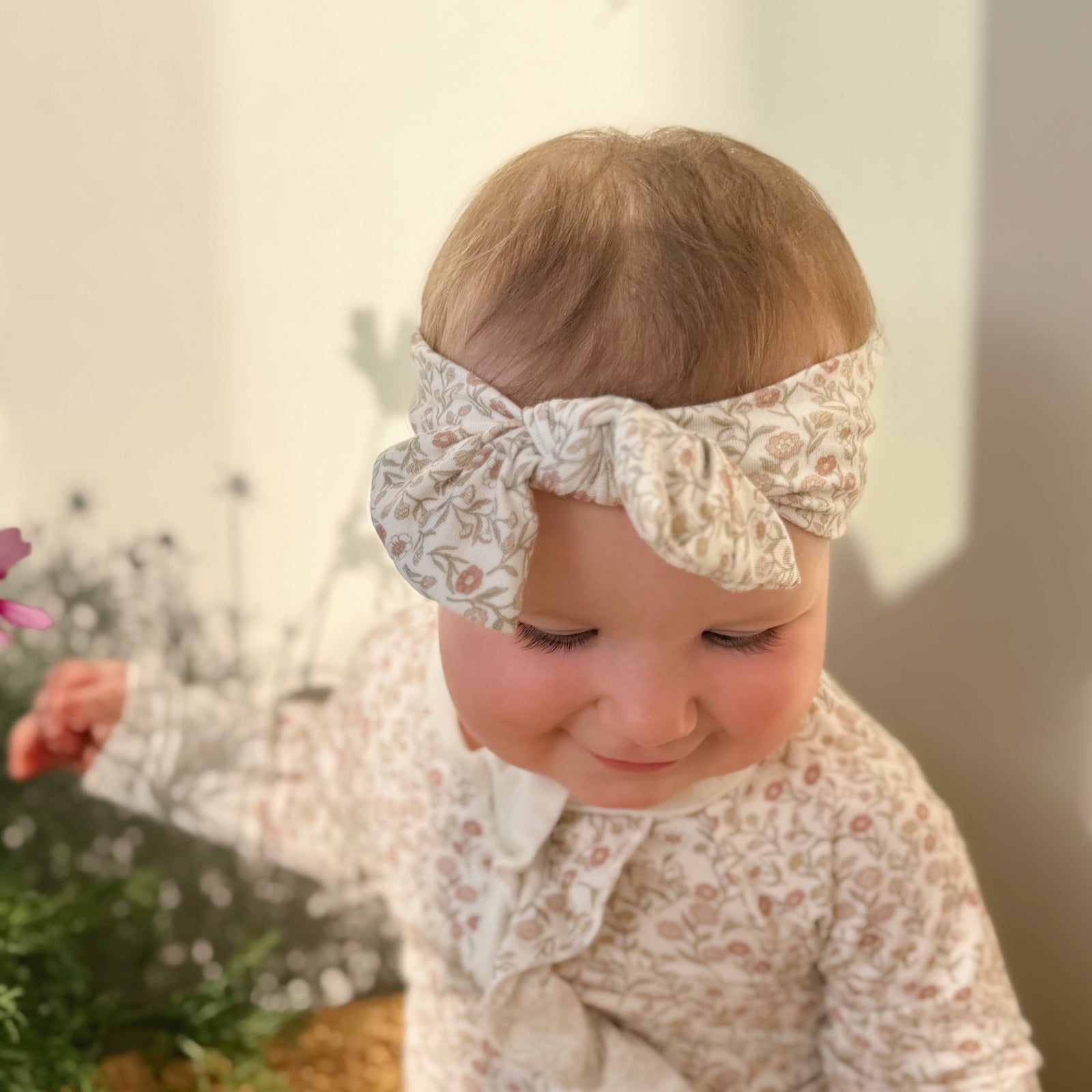 Baby wearing a floral headband and matching outfit, sitting on a couch with a blurred background