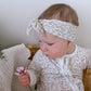 Baby wearing a floral headband and holding a flower, sitting on a textured surface.