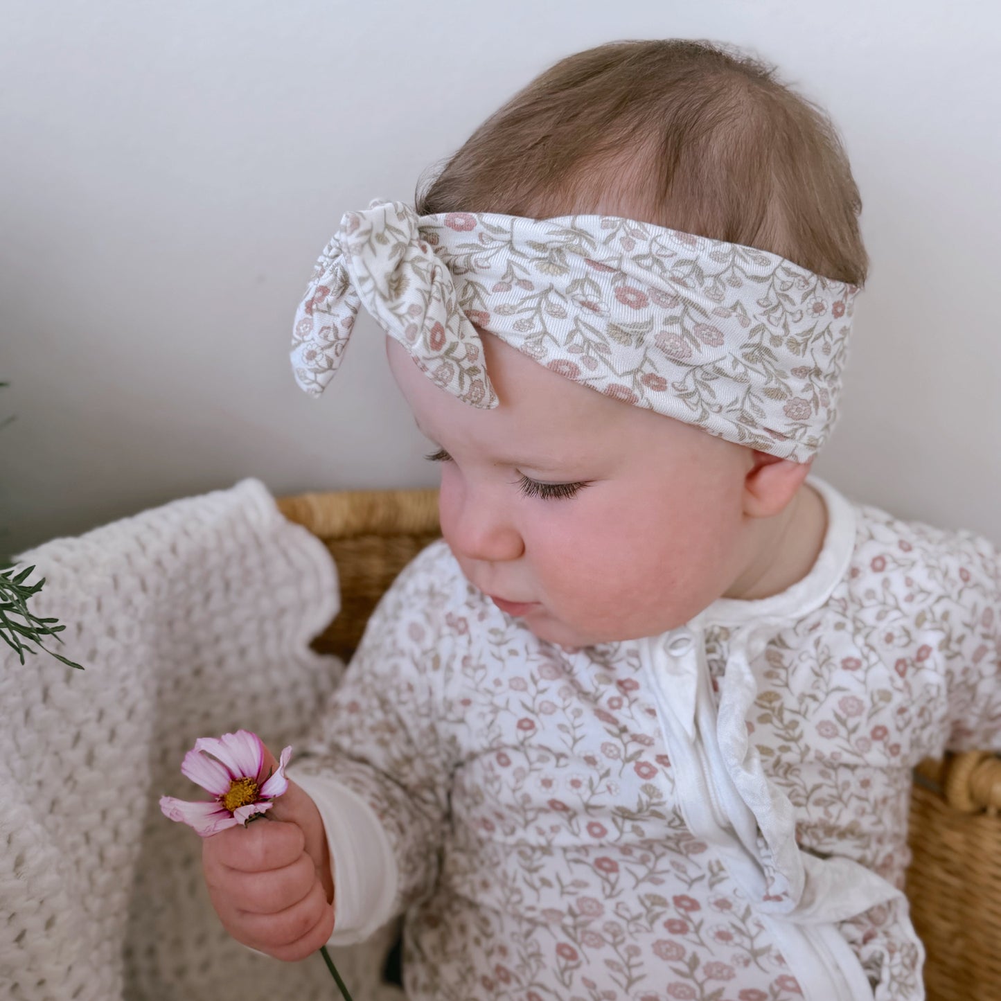Baby wearing a floral headband and holding a flower, sitting on a textured surface.