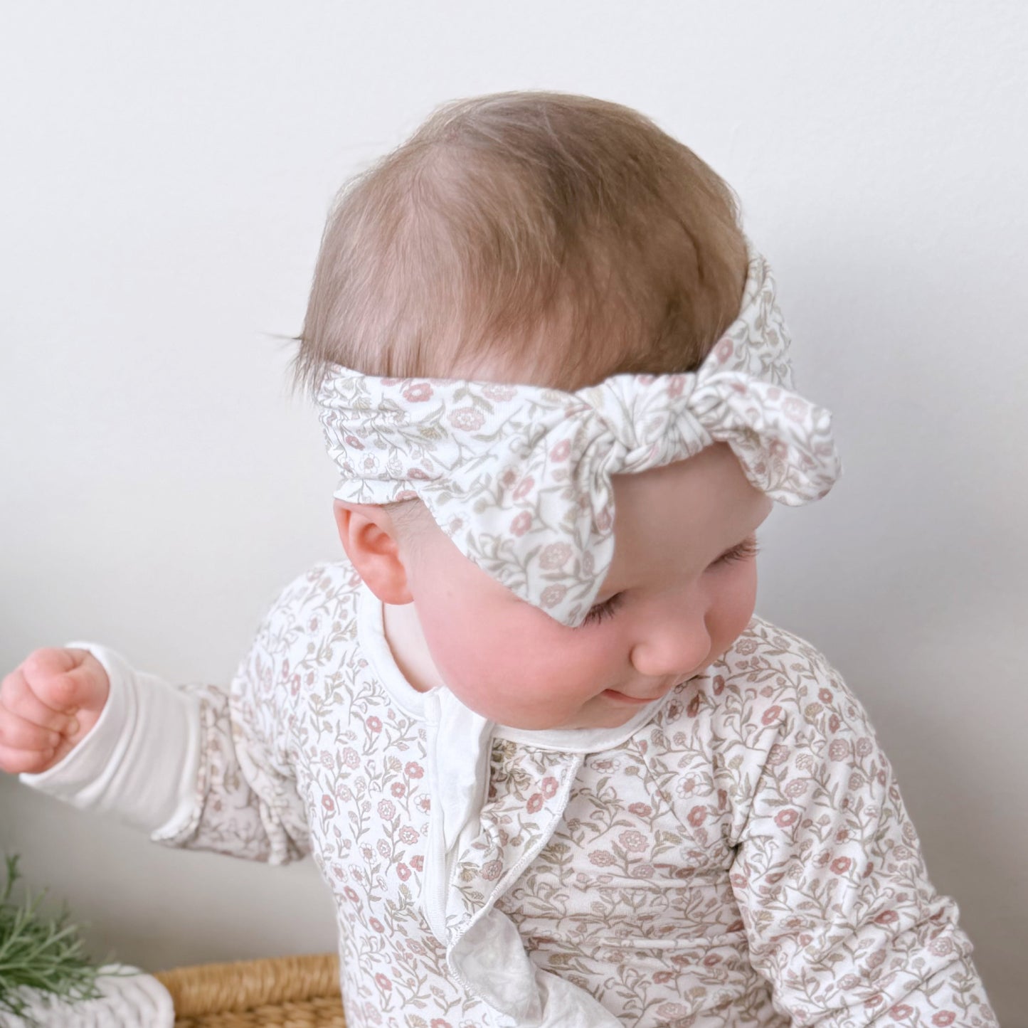 Baby in a lace outfit standing on a woven chair with a plant and flowers in the background