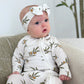 Baby wearing a white outfit with green patterns and a matching headband, sitting on a couch.