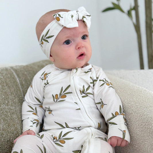 Baby wearing a white outfit with green patterns and a matching headband, sitting on a couch.